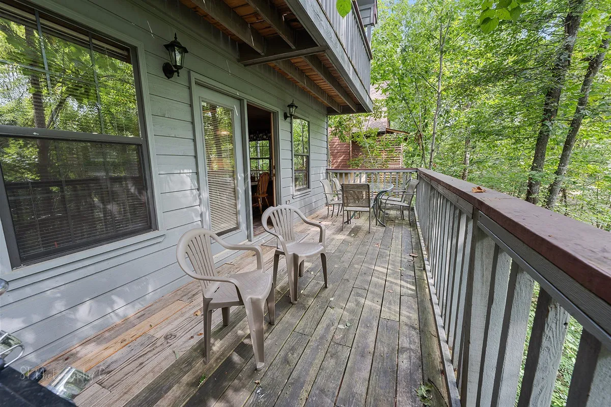 This image features a spacious wooden deck surrounded by lush trees, with plastic chairs arranged for relaxation and a glass door leading back inside. The scene is serene and inviting, providing a perfect spot for pet owners to unwind outdoors with their dogs and enjoy the natural setting.