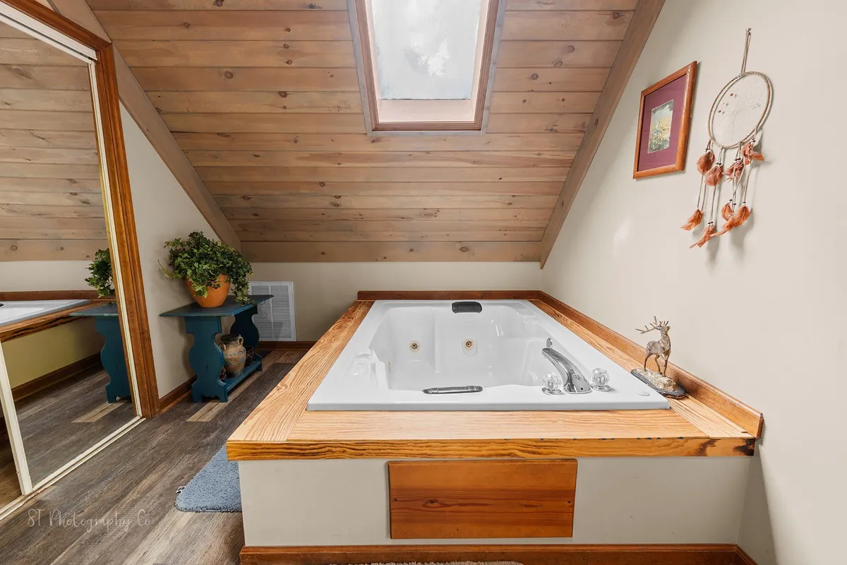 A tranquil bathroom featuring a jacuzzi tub under a skylight, wooden accents, and a decorative plant. Natural light streams in, creating a spa-like atmosphere ideal for relaxation and self-care.