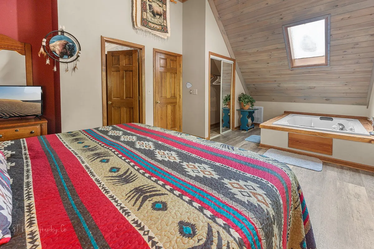 A warmly lit bedroom featuring a colorful quilted bedspread, wooden furniture, and a jacuzzi tub nestled in the corner. An angled ceiling and decorative wall hanging add character, while a TV is visible for in-room entertainment.