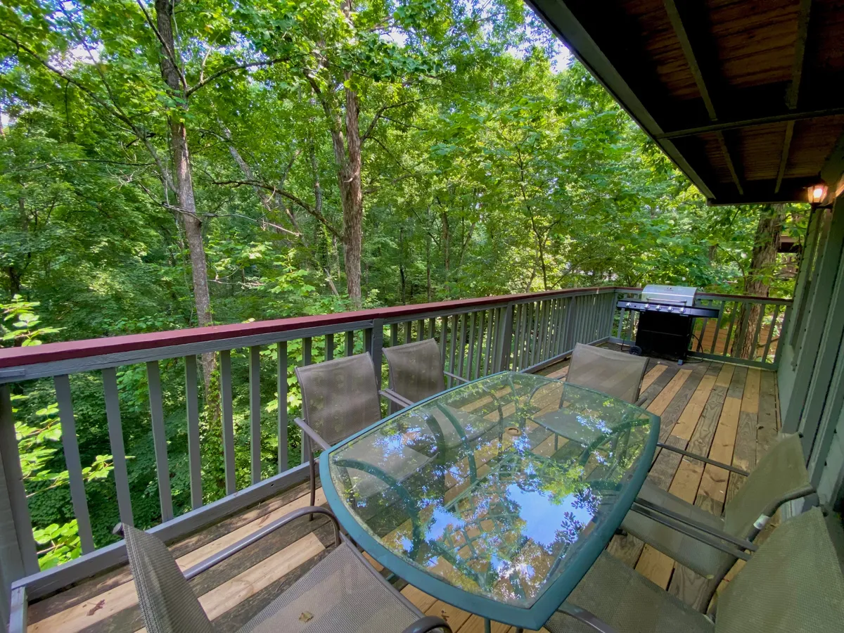 The image spotlights a wooden deck with a glass dining table, several chairs, a gas grill, and a backdrop of vibrant greenery. The grill is ready for barbecues, and the table set-up invites alfresco dining and social gatherings.