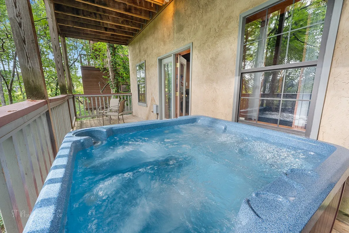 This exterior image features a wooden deck with a hot tub, large windows, and seating, all surrounded by trees. The inviting hot tub and ample deck space provide opportunities for groups to socialize, unwind, and enjoy nature views after a day of local adventures.