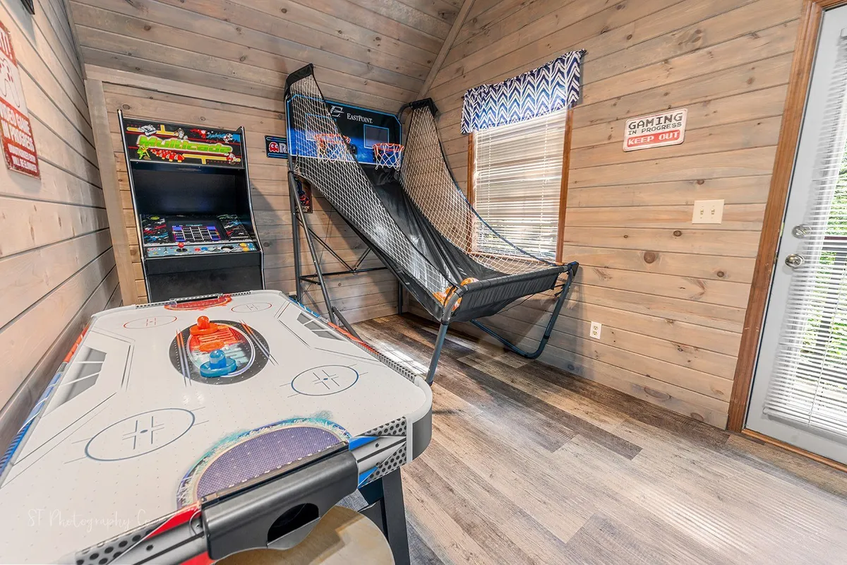 A lively game loft featuring an arcade game machine, basketball hoop, and air hockey table. Sunlight streams in from the windows, illuminating the space and highlighting the energetic, playful atmosphere. The room is set up for friendly competition and family fun, matching the section's focus on entertainment for all ages.