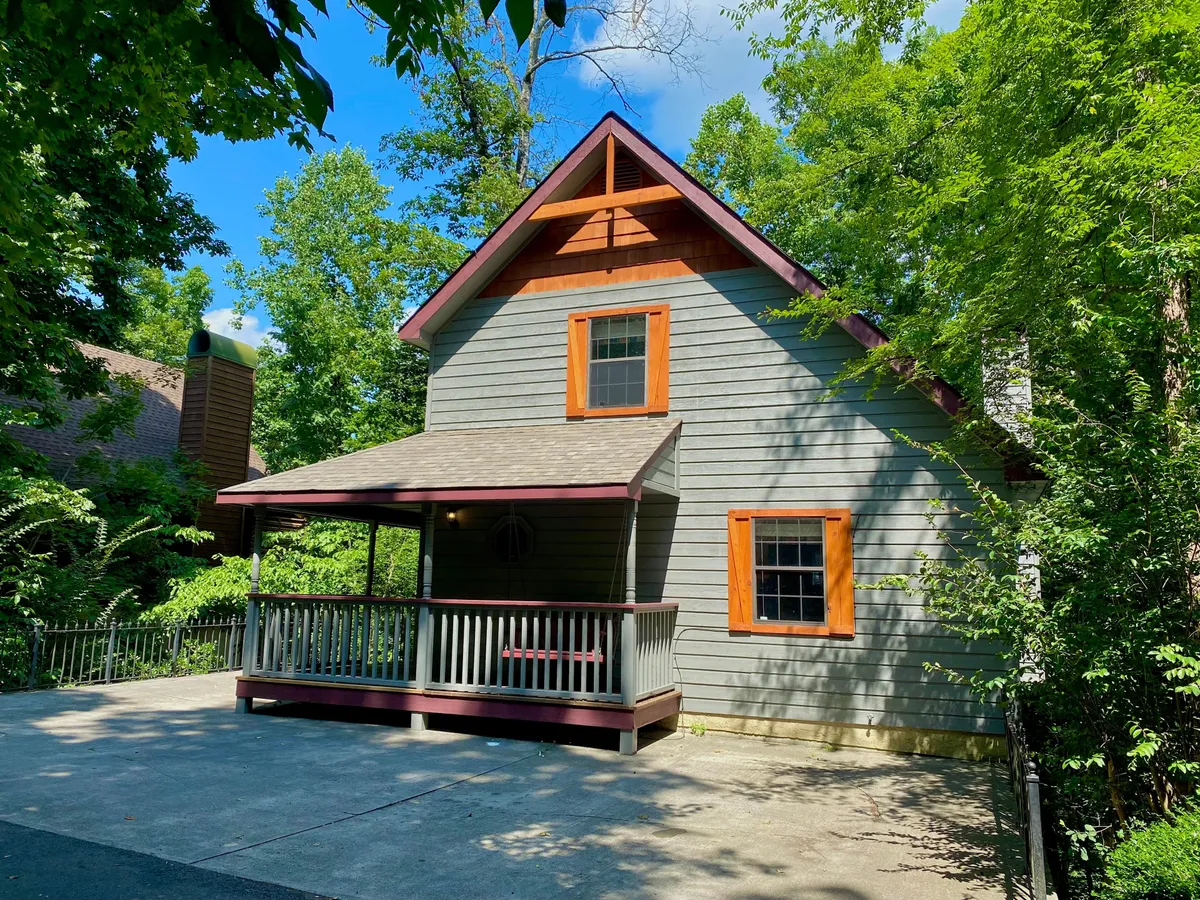 This exterior image shows the front porch of the cabin, featuring distinctive orange window trims, a sloping roof, and cozy seating. The setting is surrounded by nature, evoking the mountain atmosphere and inviting families to relax outdoors or gather before heading out to explore local attractions.