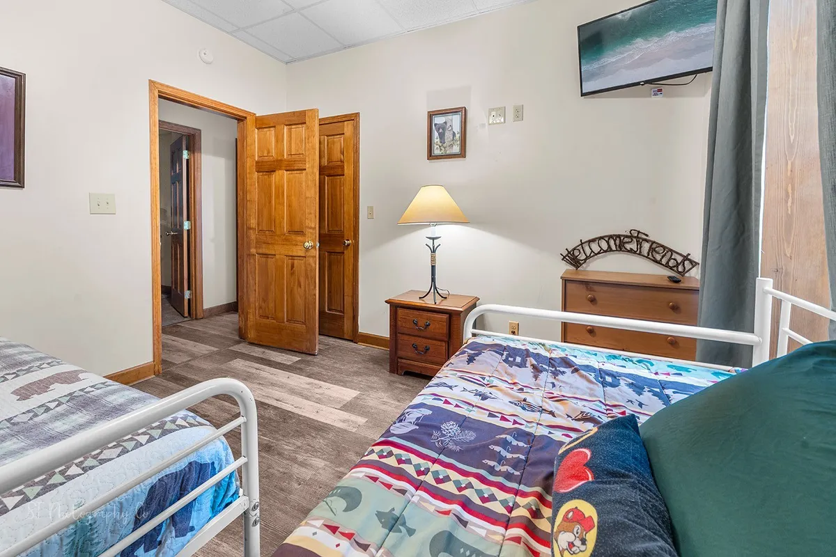 A room featuring two beds, a nightstand with a lamp, an open door, and a wall-mounted television. The setup suggests a welcoming space for multiple guests, ideal for families or kids. The lighting is soft and warm.