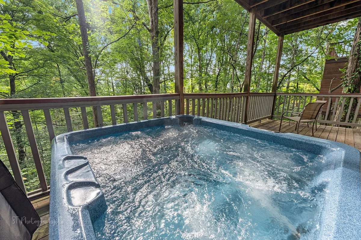 This image captures a serene outdoor deck area featuring a bubbling hot tub framed by wooden deck railings, surrounded by lush greenery. The hot tub is the focal point, inviting guests to relax while enjoying the mountain air and tranquil forest setting.