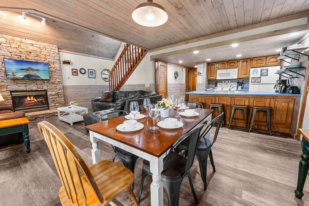 An open-concept area connecting a dining table set for six, a comfortable seating area, a stone fireplace, and a fully equipped kitchen with wooden cabinetry. The scene is warm, inviting, and ideal for shared meals or entertaining guests.