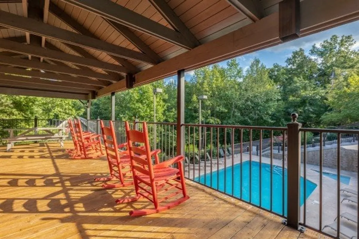 This exterior image showcases a sparkling swimming pool bordered by a wooden deck, with inviting red rocking chairs positioned for relaxation. The scene captures both the tranquil atmosphere and the opportunity for summer fun, with lush greenery providing a picturesque backdrop.